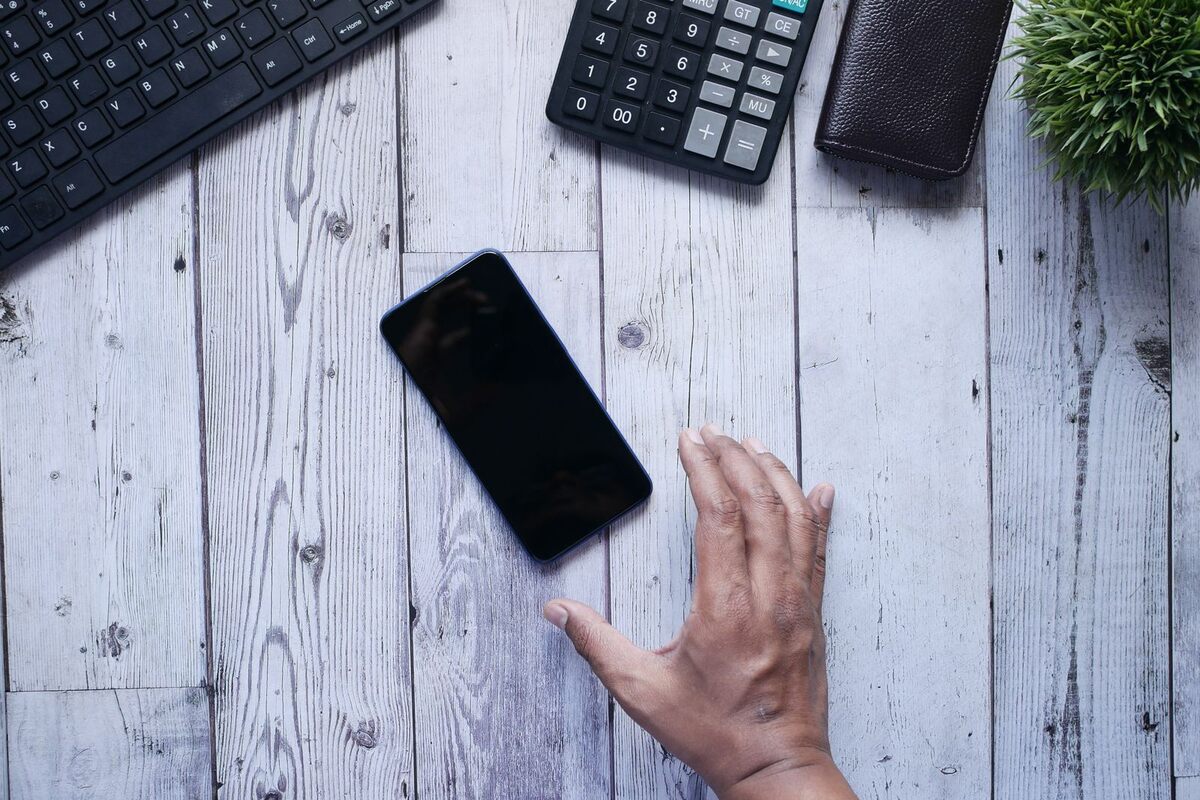 Illustration: a smartphone lying on a wooden desk beside a keyboard, calculator and wallet, with a hand reaching to pick it up.
